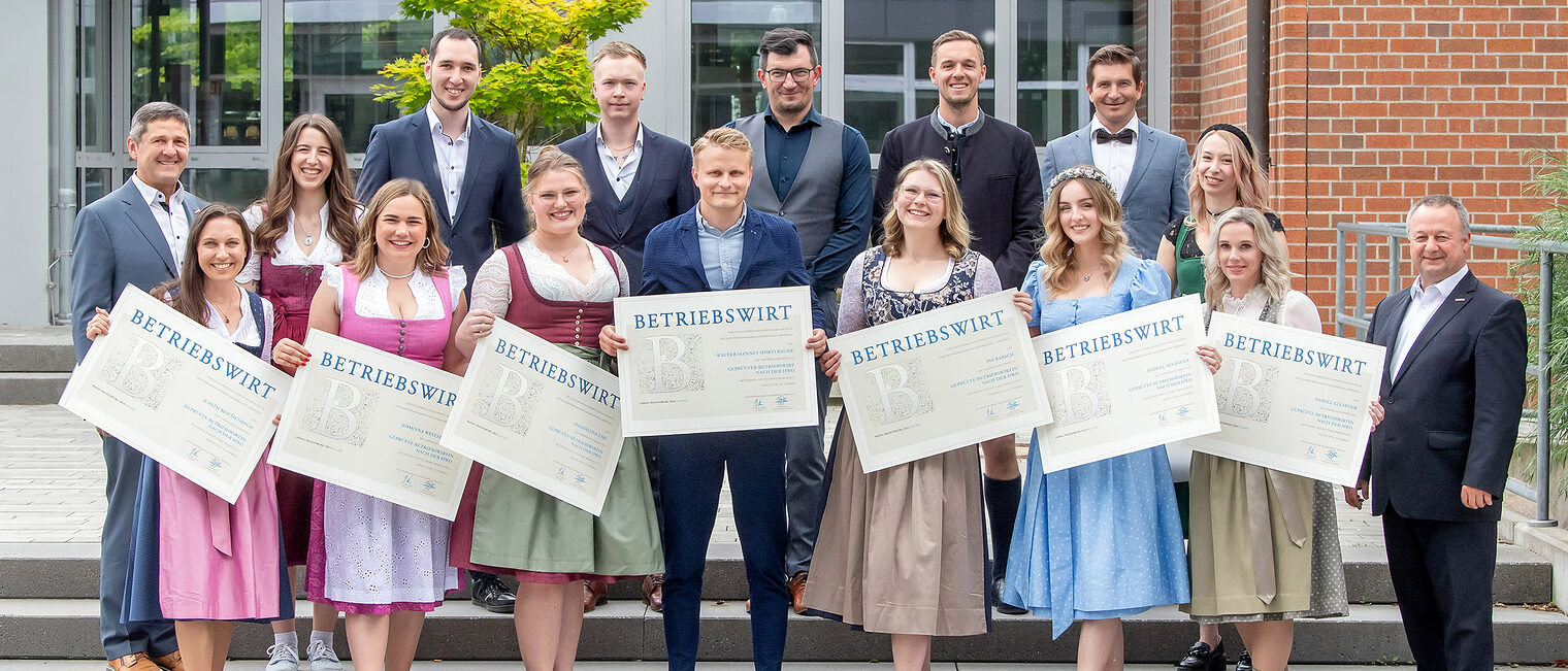 Vizepräsident Christian Läpple und Fachbereichsleiter Unternehmensführung Manfred Wellner übergaben die Zeugnisse an die Betriebswirte. Die Betriebswirte halten fröhlich ihre Zeugnisse in die Kamera. Die Ehrengäste freuen sich mit ihnen.